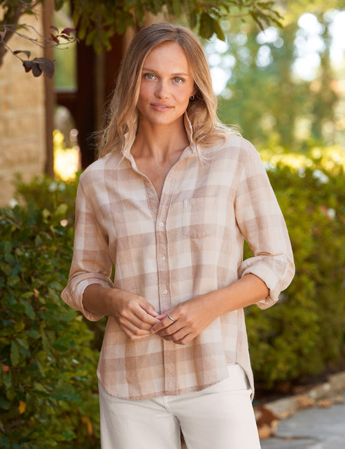 A woman wearing a light beige and cream plaid button-down shirt with rolled-up sleeves, standing outdoors in front of a blurred garden background., front view