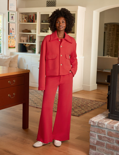 A woman stands in a living room wearing a bright red two-piece suit consisting of a cropped jacket with front flap pockets and matching wide-leg pants. She is wearing white slip-on shoes., front view