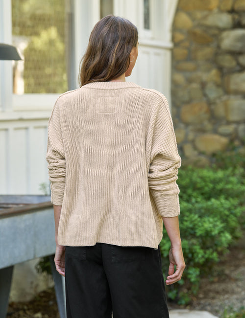 Person wearing a beige sweater and black pants standing outdoors., alternate view