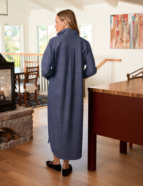 A woman wearing a long blue denim shirt dress, viewed from behind in a bright, modern dining room with wood floors and a fireplace., alternate view