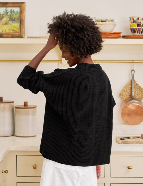 Woman standing in a kitchen wearing a black sweater and white skirt., alternate view