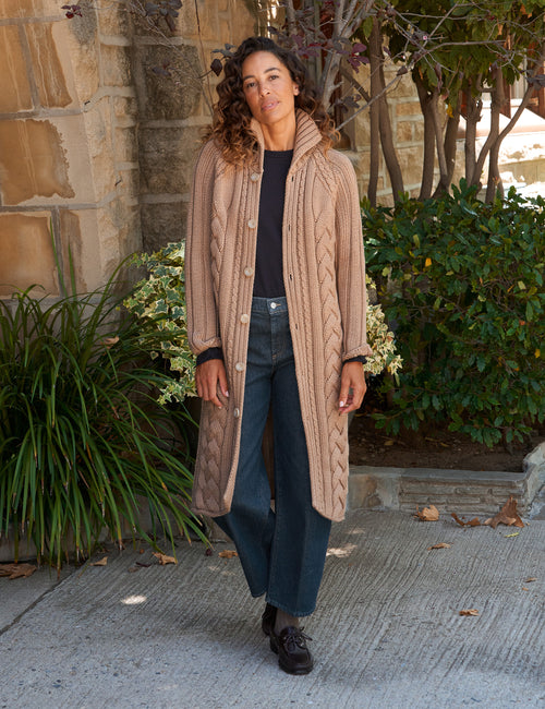 A woman stands outdoors wearing a long beige cable-knit cardigan over a black top and dark blue wide-leg jeans, with dark loafers, in front of a stone wall and green plants., front view