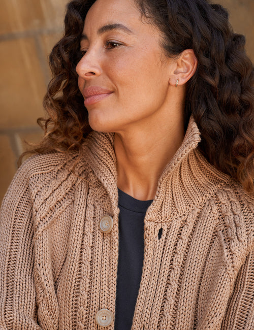 A close-up of a woman wearing a chunky knit cardigan in a warm beige/tan color over a dark shirt, with a blurred stone wall background., view 3
