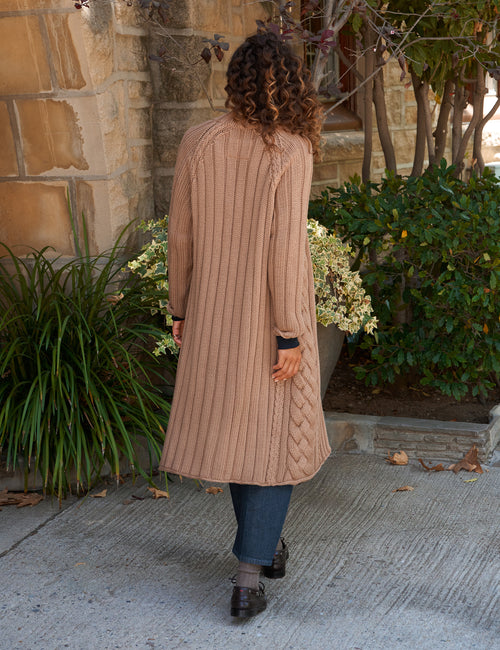 A woman viewed from behind wearing a long, beige cable-knit cardigan with blue jeans and dark loafers, standing on a concrete sidewalk in front of a stone building with green plants., alternate view