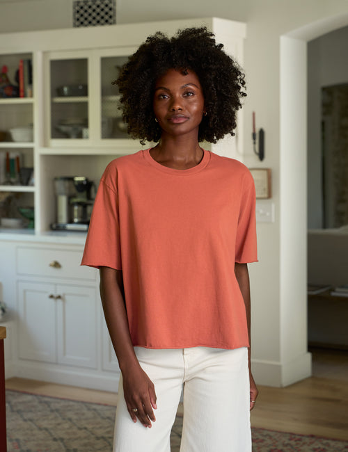 A woman with curly black hair wearing a muted orange short-sleeve crewneck t-shirt and white pants, standing in a bright, modern kitchen., front view