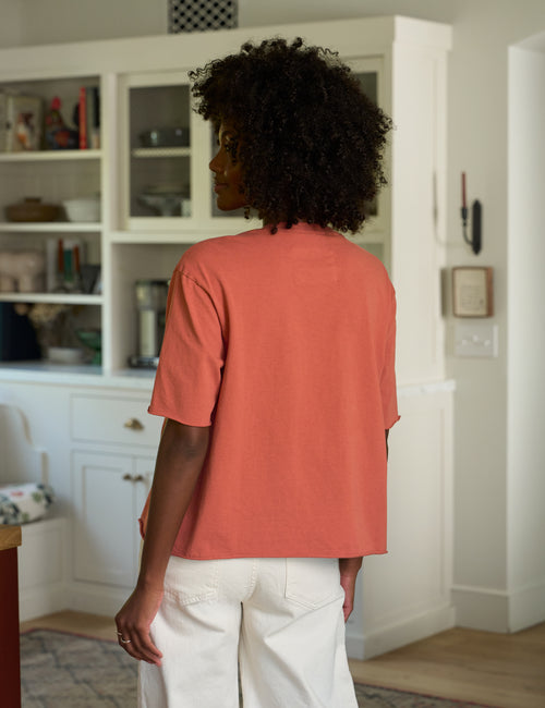 A woman with curly dark hair is shown from behind wearing a orange short-sleeve t-shirt and white pants in a bright, modern home interior with white cabinetry., alternate view