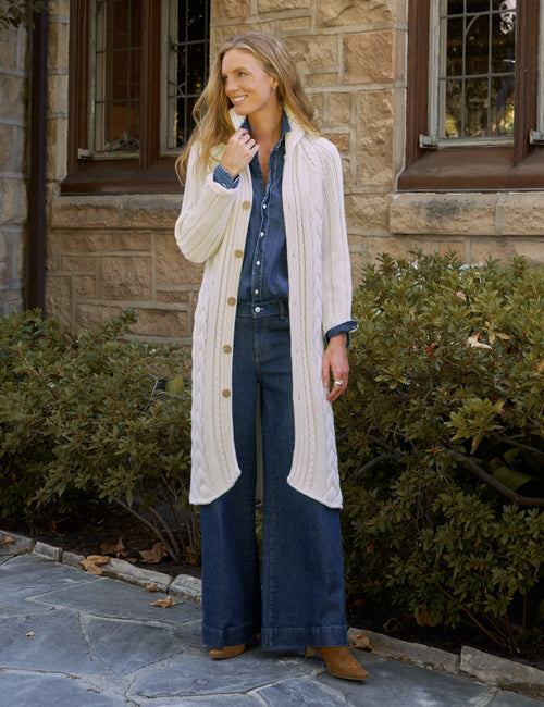 A woman stands outdoors in front of a stone building and green shrubs, wearing a long cream cable-knit cardigan over a blue button-up shirt and dark blue wide-leg jeans, with tan ankle boots., front view