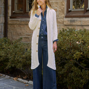 A woman stands outdoors in front of a stone building and green shrubs, wearing a long cream cable-knit cardigan over a blue button-up shirt and dark blue wide-leg jeans, with tan ankle boots.