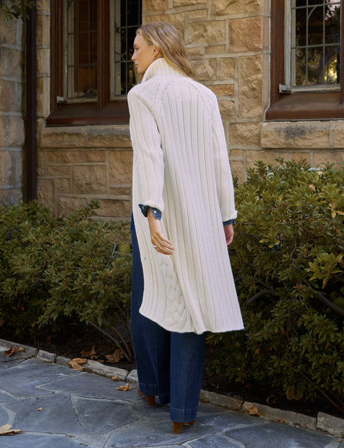 A woman stands outdoors in front of a stone building, wearing a long cream cable-knit stand collar cardigan over a blue denim shirt and blue jeans., alternate view