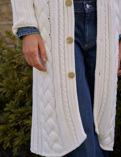 A close-up of a person wearing a long cream cable-knit cardigan with large tan buttons, layered over a denim shirt and paired with blue jeans, with green foliage in the background., view 4