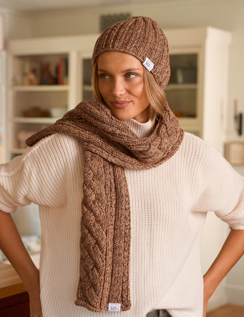 A woman wearing a chunky brown cable-knit beanie, matching scarf, and cream-colored ribbed sweater, standing in a softly lit indoor room., front view