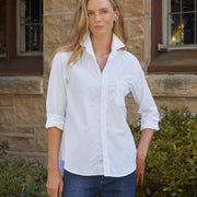 A woman wearing a white long-sleeve button-down shirt with rolled-up sleeves and blue jeans, standing outdoors in front of a stone building with green bushes.