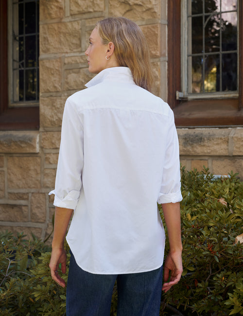 A woman wearing a white long-sleeve button-down shirt with rolled-up sleeves and dark blue jeans, standing outdoors in front of a stone building., alternate view