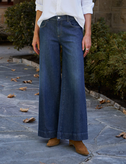 A person wearing dark blue wide-leg denim jeans, a white button-up shirt, and tan suede boots, standing on a stone patio with greenery in the background., front view