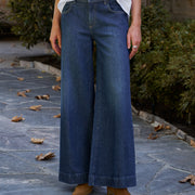 A person wearing dark blue wide-leg denim jeans, a white button-up shirt, and tan suede boots, standing on a stone patio with greenery in the background.