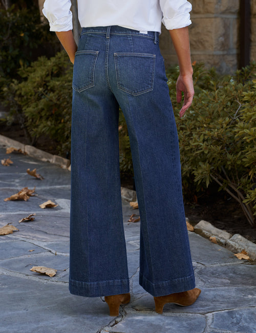 A person shown from the back wearing dark blue wide-leg denim jeans, a white long-sleeve blouse, and brown ankle boots on a stone walkway with green shrubs in the background., alternate view