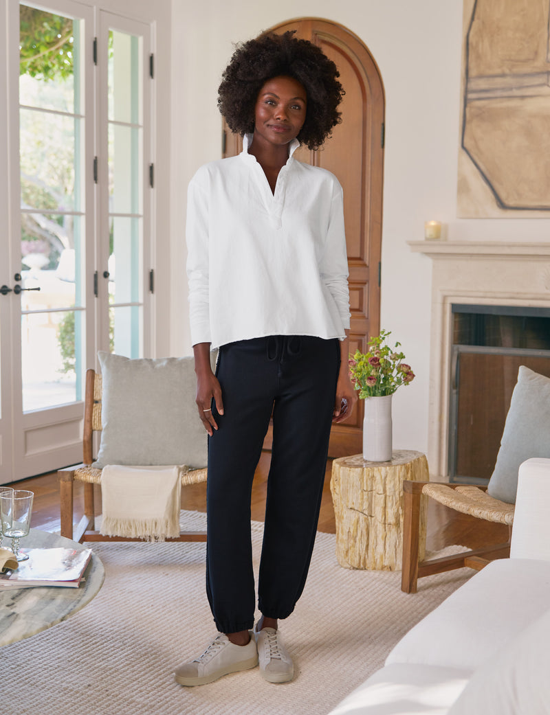 Woman standing in a living room wearing a white shirt and navy pants.