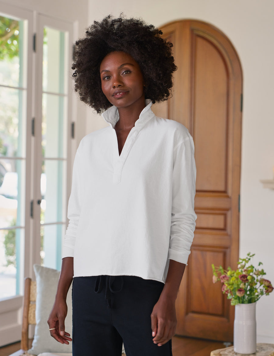 Woman wearing a white long-sleeve shirt standing in a room with a wooden door and a vase of flowers.