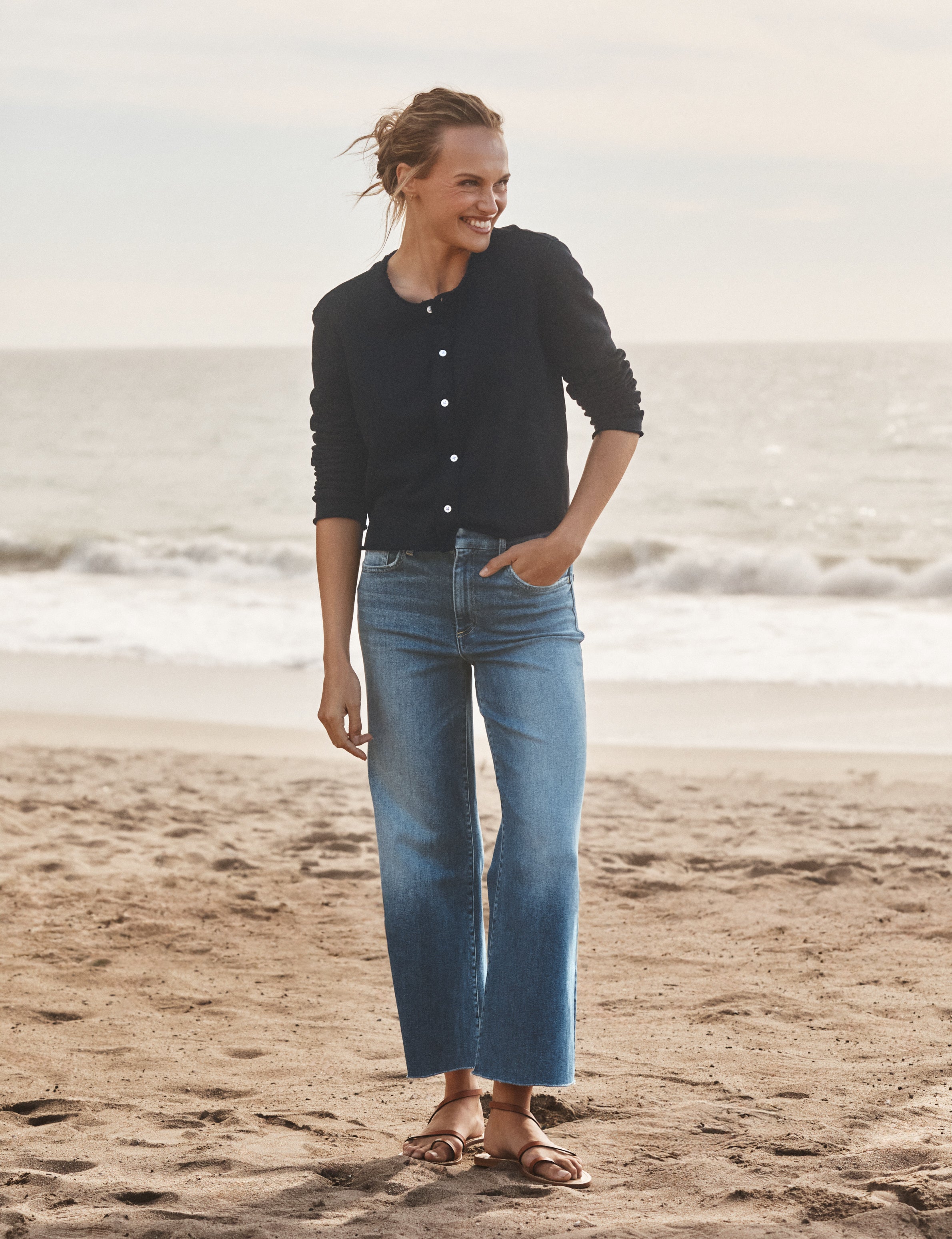Woman standing on a beach wearing a navy cardigan and blue jeans., view 4