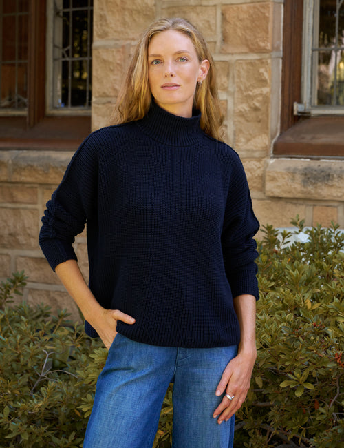 A woman wearing a dark navy ribbed knit mock neck sweater and blue jeans, standing outdoors in front of a stone building., front view