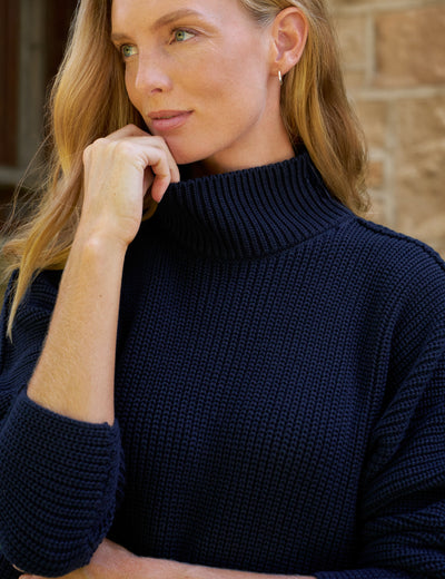 A close-up of a woman wearing a dark navy blue ribbed knit turtleneck sweater, resting her chin on her hand against a blurred stone wall background.