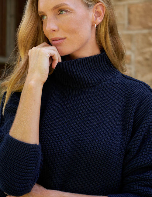 A close-up of a woman wearing a dark navy blue ribbed knit turtleneck sweater, resting her chin on her hand against a blurred stone wall background., view 3