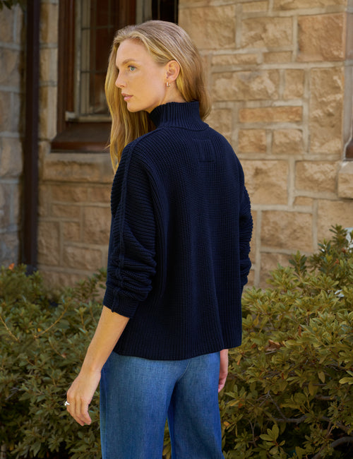 A woman wearing a navy blue ribbed turtleneck sweater and blue jeans, standing outdoors in front of a stone wall and green bushes., alternate view