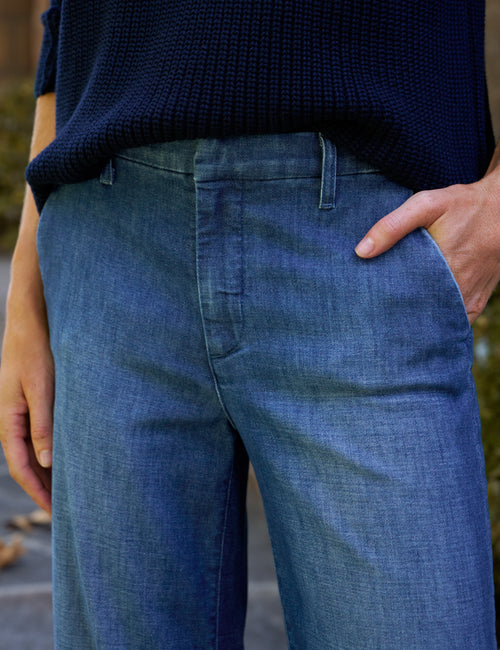 Close-up of a person wearing blue denim pants with a dark navy ribbed knit top, standing outdoors with one hand in the pocket., view 3