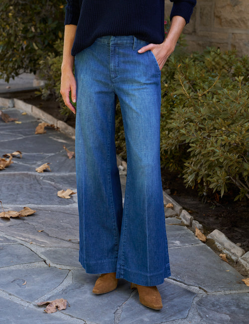 A person wearing blue wide-leg denim jeans, a dark navy ribbed sweater, and tan ankle boots, standing on a stone patio with green shrubs in the background., front view