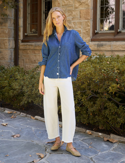 A woman stands outdoors on a stone walkway in front of a stone building, wearing a blue denim button-up shirt, off-white wide-leg cropped pants, and tan flats., view 5