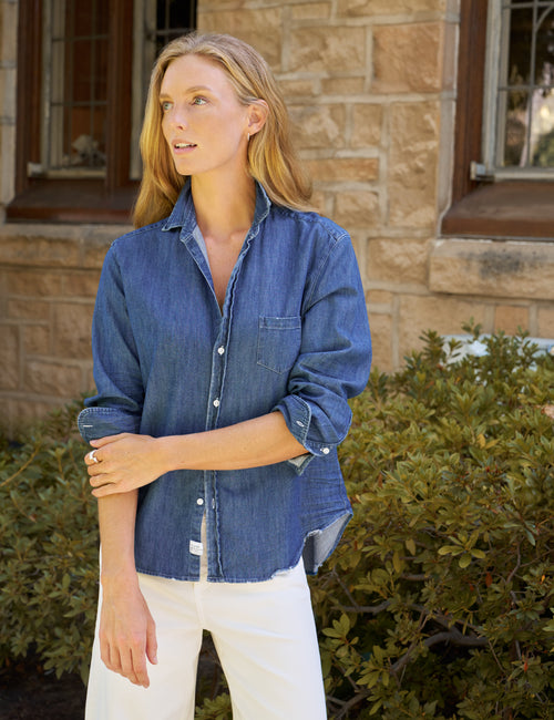 A woman wearing a blue denim button-up shirt and white pants stands outdoors in front of a stone building and green bushes., view 4