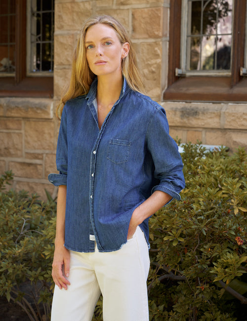A woman wearing a blue denim button-up shirt with rolled sleeves and white pants, standing outdoors in front of a stone building and green bushes., front view