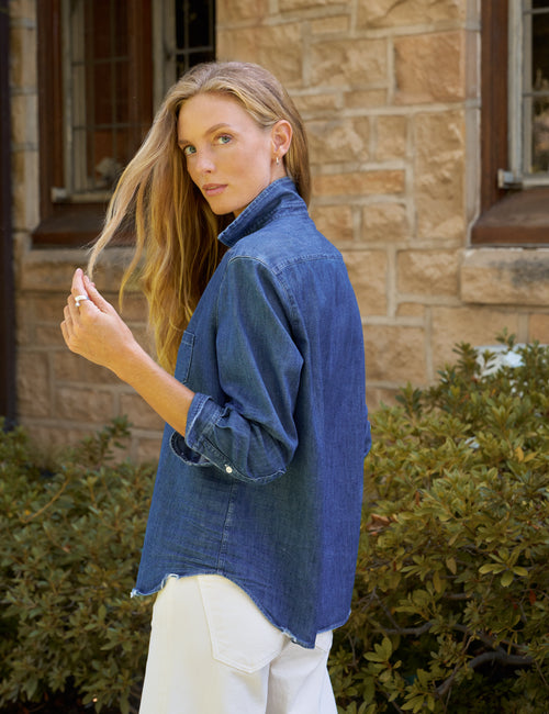 A woman wearing a blue denim button-up shirt and white pants, standing outdoors in front of a stone building with green bushes., alternate view