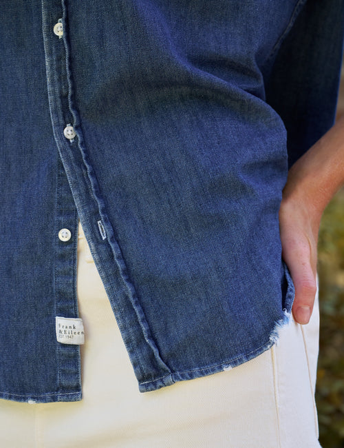 Close-up of a person wearing a dark blue denim button-up shirt with white buttons and off-white pants, with a hand in the shirt pocket., view 3