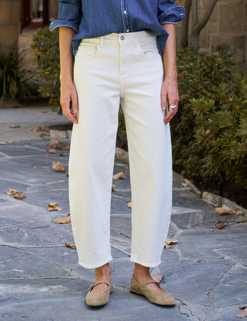A person wearing off-white cropped jeans, a blue button-up shirt, and beige flats, standing on a stone walkway outdoors., front view