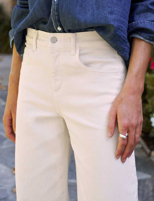 A close-up of a person wearing off-white or cream-colored barrel-leg jeans with a blue denim shirt tucked into them. The photo is cropped from the chest down, showing the front pocket, belt loops, button, and the person's hand resting on their thigh., view 3