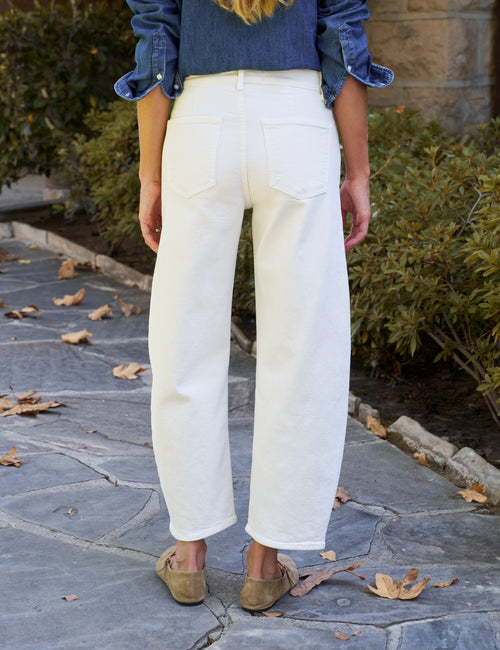 A person shown from the back wearing off-white barrel-leg cropped jeans, a blue denim shirt, and tan slip-on shoes on a stone patio with green bushes in the background., alternate view