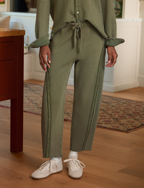 A person wearing an olive green textured knit two-piece set with matching drawstring tie waist and matching long-sleeve top, paired with white sneakers in a home interior setting., front view