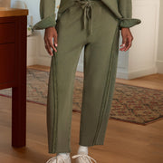 A person wearing an olive green textured knit two-piece set with matching drawstring tie waist and matching long-sleeve top, paired with white sneakers in a home interior setting.