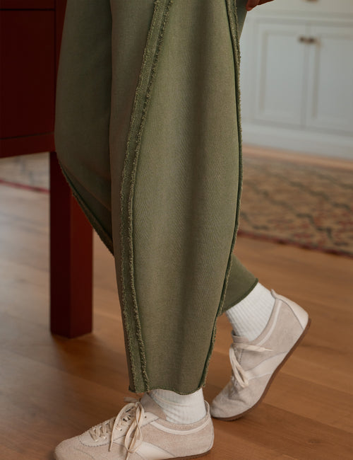 A close-up lifestyle shot of a person wearing olive green textured knit pants with raw, frayed edges, styled with white crew socks and beige sneakers in a warm indoor setting with a wooden floor., view 3