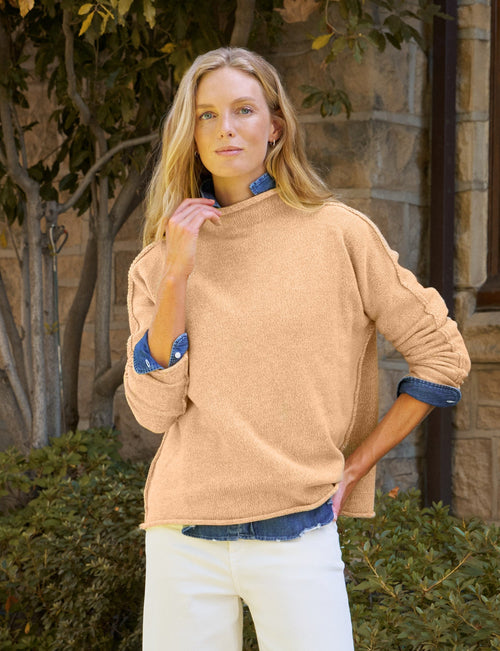 A woman stands outdoors on a stone walkway in front of a stone building, wearing a beige knit sweater over a blue collared shirt, white cropped pants, and tan loafers., front view