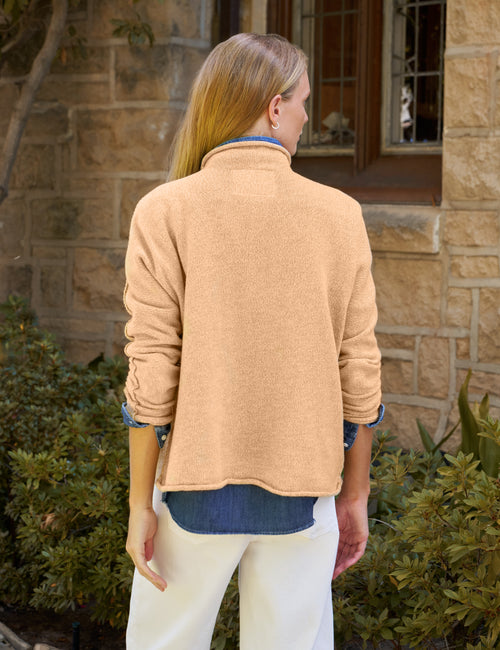 A woman viewed from behind wearing a light peach-beige knit quarter-zip fleece jacket over a blue shirt and white pants, standing outdoors in front of a stone building., alternate view