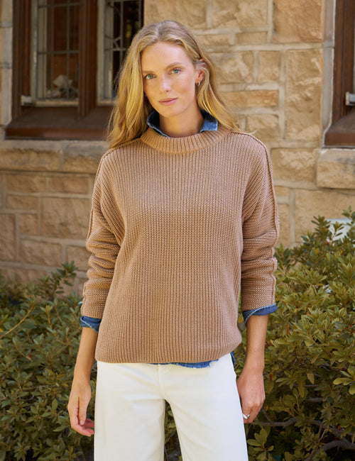 A woman wearing a ribbed knit crewneck sweater in camel brown, layered over a blue collared shirt and paired with white pants, standing outdoors in front of a stone building., front view