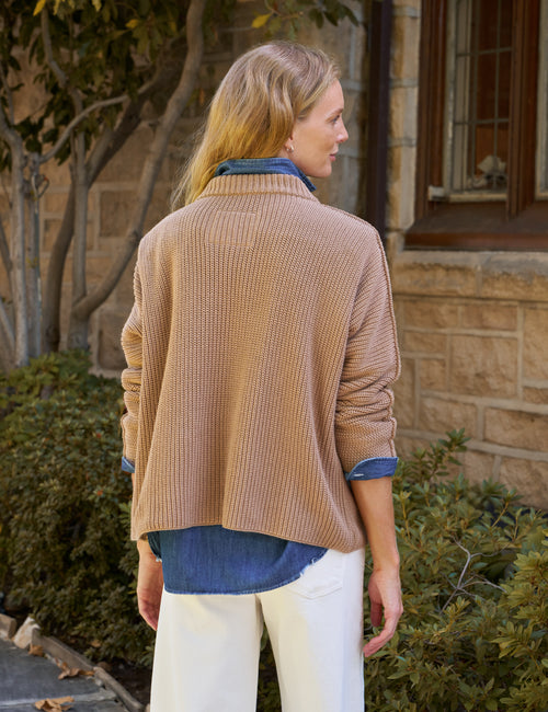 A woman shown from the back wearing a beige ribbed knit cardigan over a blue denim shirt and white pants, standing outdoors in front of a stone building., alternate view