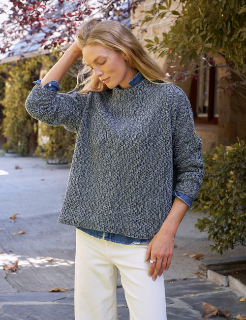 A woman wearing a textured gray knit mock-neck sweater with rolled-up sleeves showing a blue denim lining, paired with white pants, standing outdoors on a stone path., front view