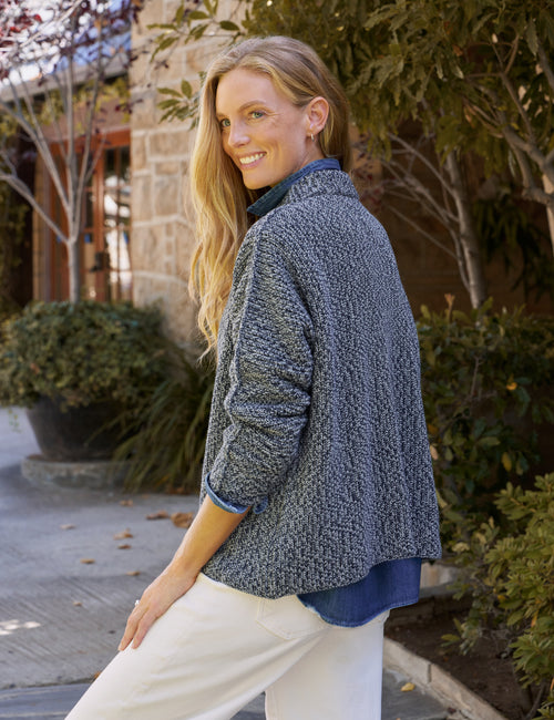 A woman wearing a textured blue and gray sweater over a dark blue top and white pants, standing outdoors in front of a stone building and greenery., alternate view