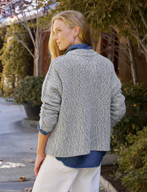 A woman wearing a textured gray knit cardigan over a blue shirt and white pants, photographed outdoors on a paved walkway with trees and shrubs in the background., alternate view