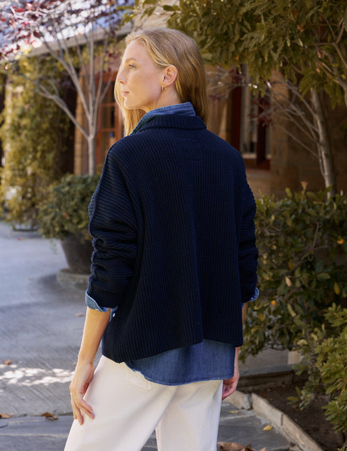A woman wearing a navy blue ribbed knit sweater over a blue shirt and white pants, standing outdoors on a paved walkway with trees and a building in the background., alternate view