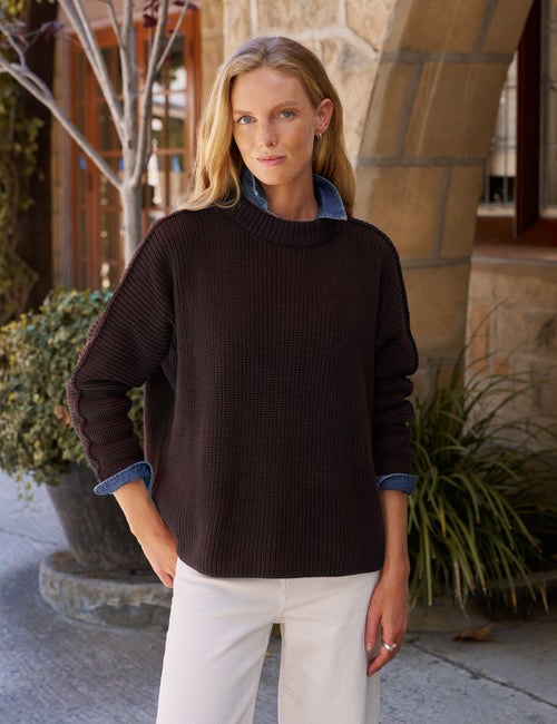 A woman wearing a dark brown ribbed knit sweater over a blue collared shirt, paired with white pants, standing outdoors in front of a stone building., front view