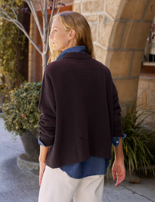 A woman wearing a dark brown ribbed knit cardigan over a blue denim shirt and cream pants, standing outdoors in front of a stone building., alternate view